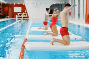 Adults performing balance exercises on floating boards in indoor swimming pool.